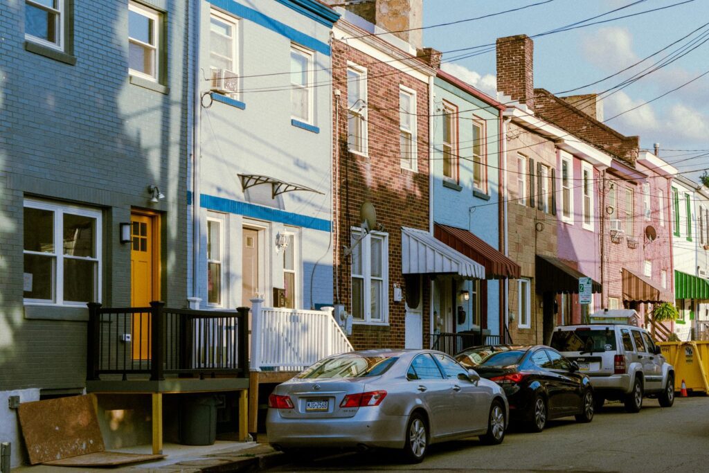 A row of attached two story brick row houses in blue, pink and tan with awnings and parked cars along the street.
