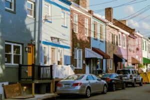 A row of attached two story brick row houses in blue, pink and tan with awnings and parked cars along the street.