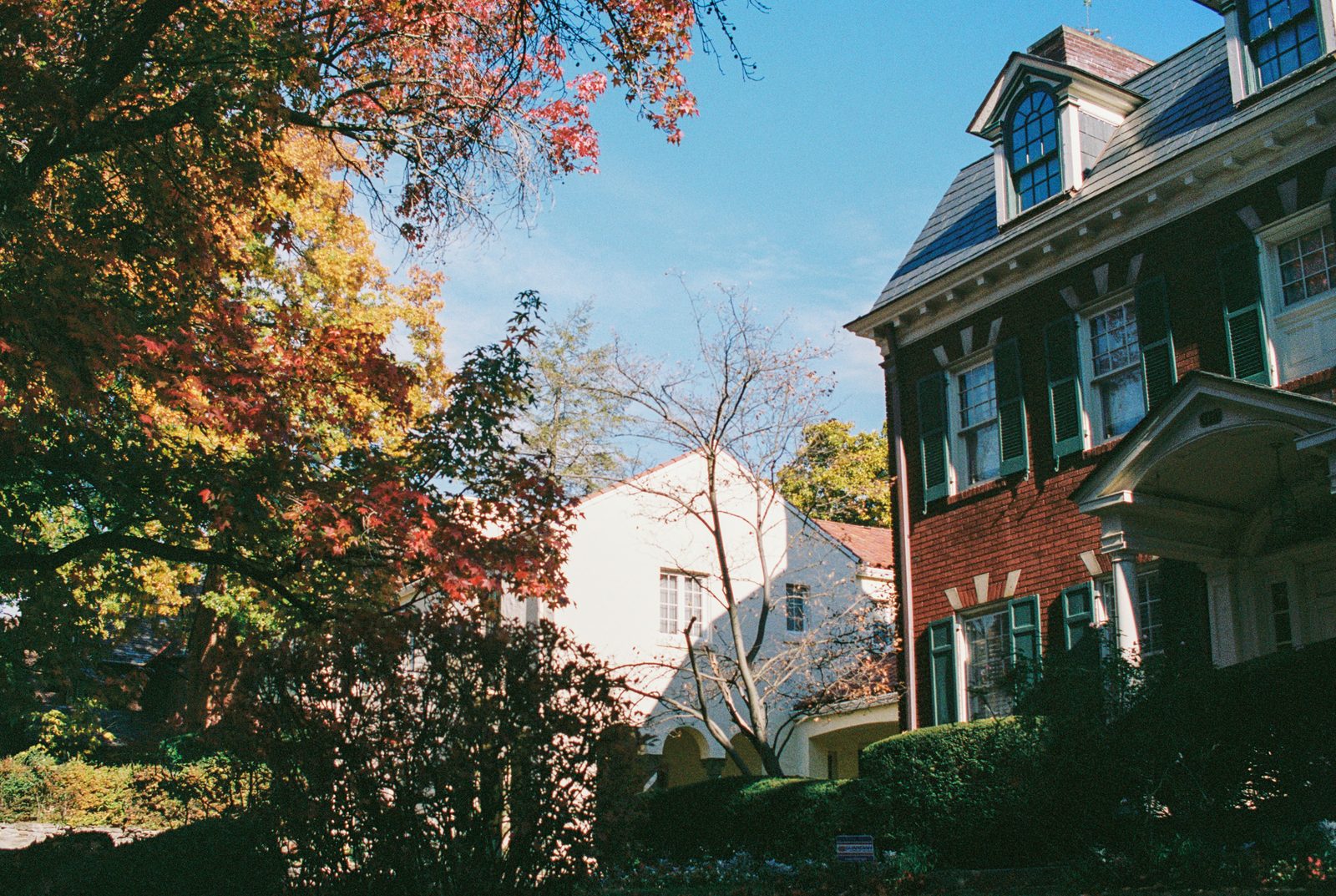 A two and a half story brick house with white trim and dormers among colorful autumn trees.