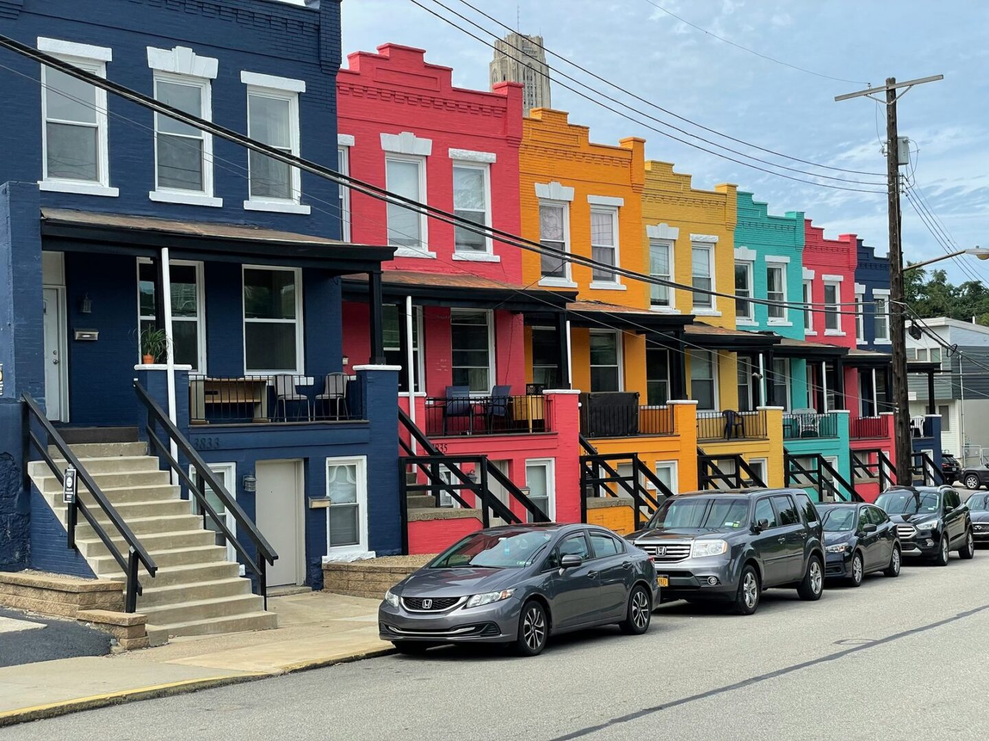 A row of brightly painted attached houses in blue, red, orange and teal with front porches and parked cars.