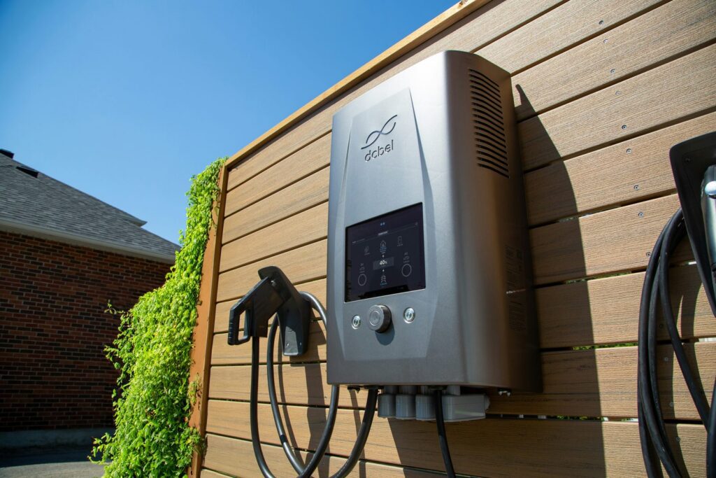 A silver dcbel home EV charger with a screen mounted on a wood fence outdoors, J1772 cable holstered beside it, brick houses in the background.