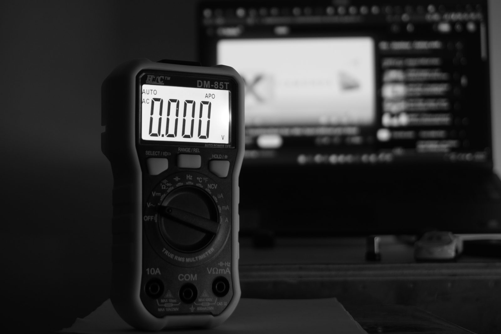 A gray digital multimeter standing on a dark surface with its display lit.