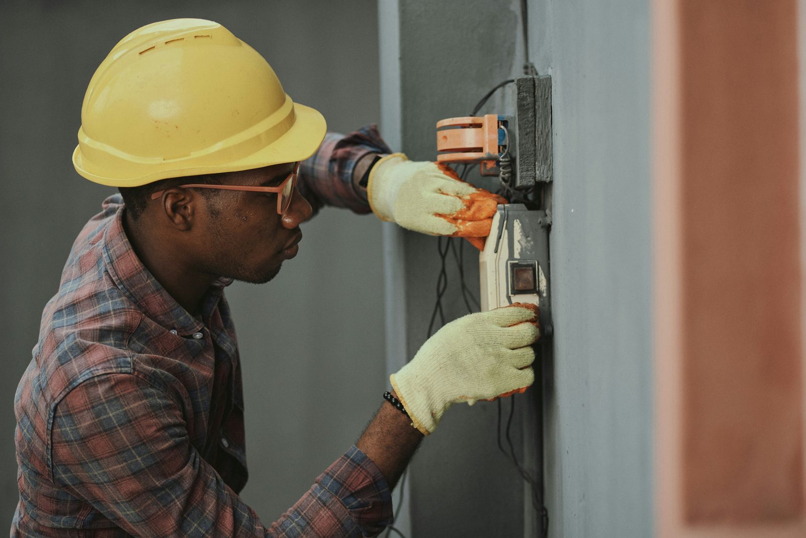 An electrician in a yellow hard hat and gloves works on a small gray electrical box on a gray exterior wall.