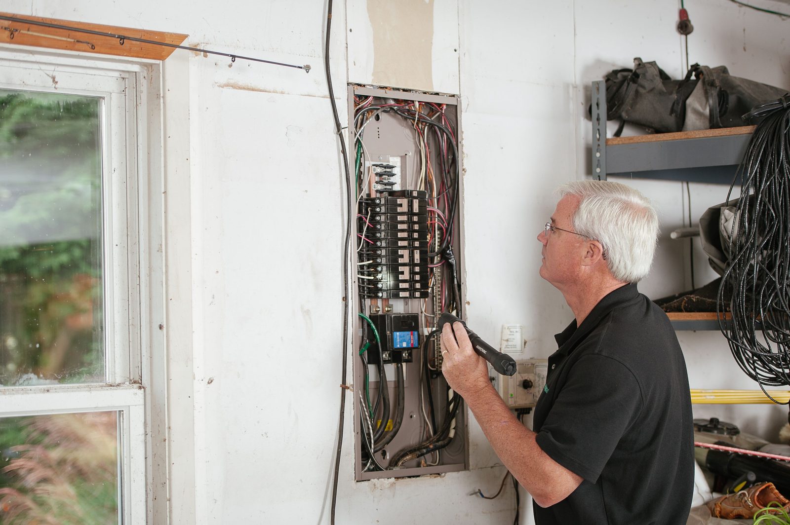 A gray haired electrician in a black polo examines an open breaker panel on a white basement wall with a flashlight.