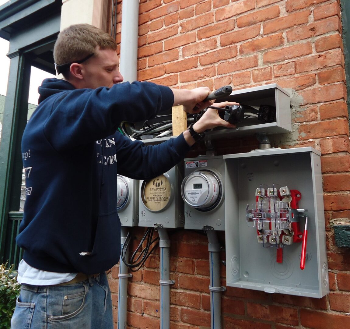 An electrician installs wiring into new meter sockets and an open disconnect on the brick wall of a multifamily house.