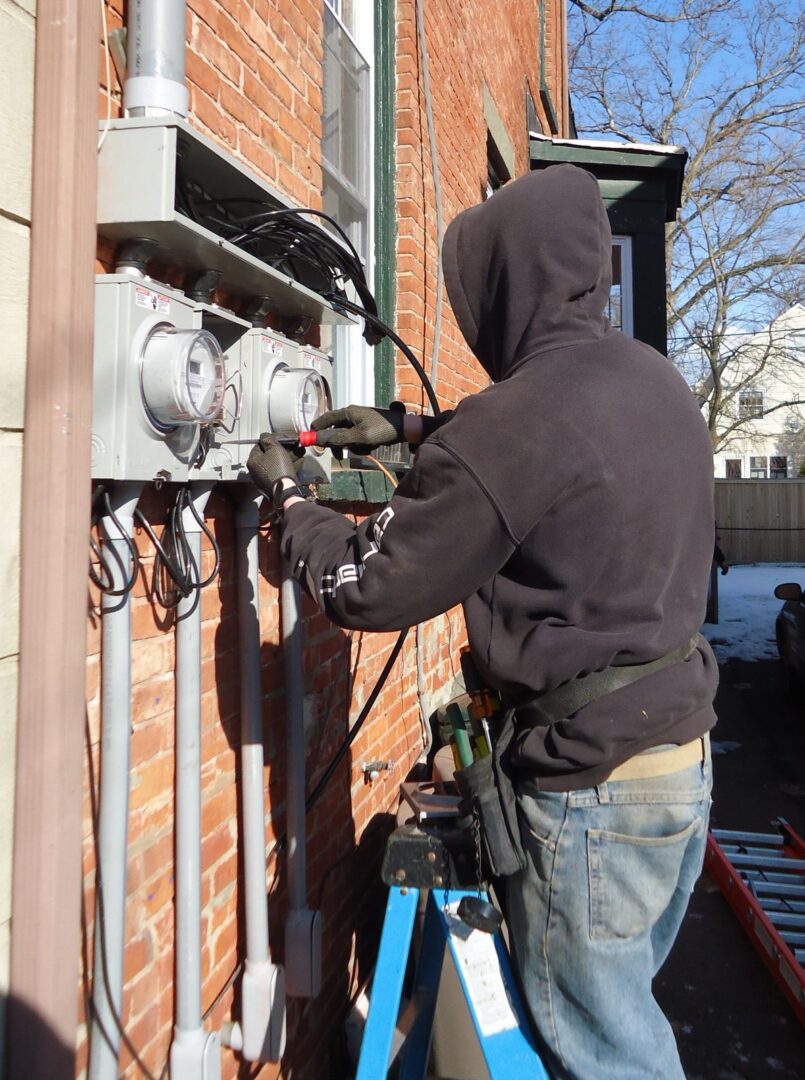 An electrician in a hooded sweatshirt on a ladder wires a row of new meter sockets on the brick wall of a house in winter.