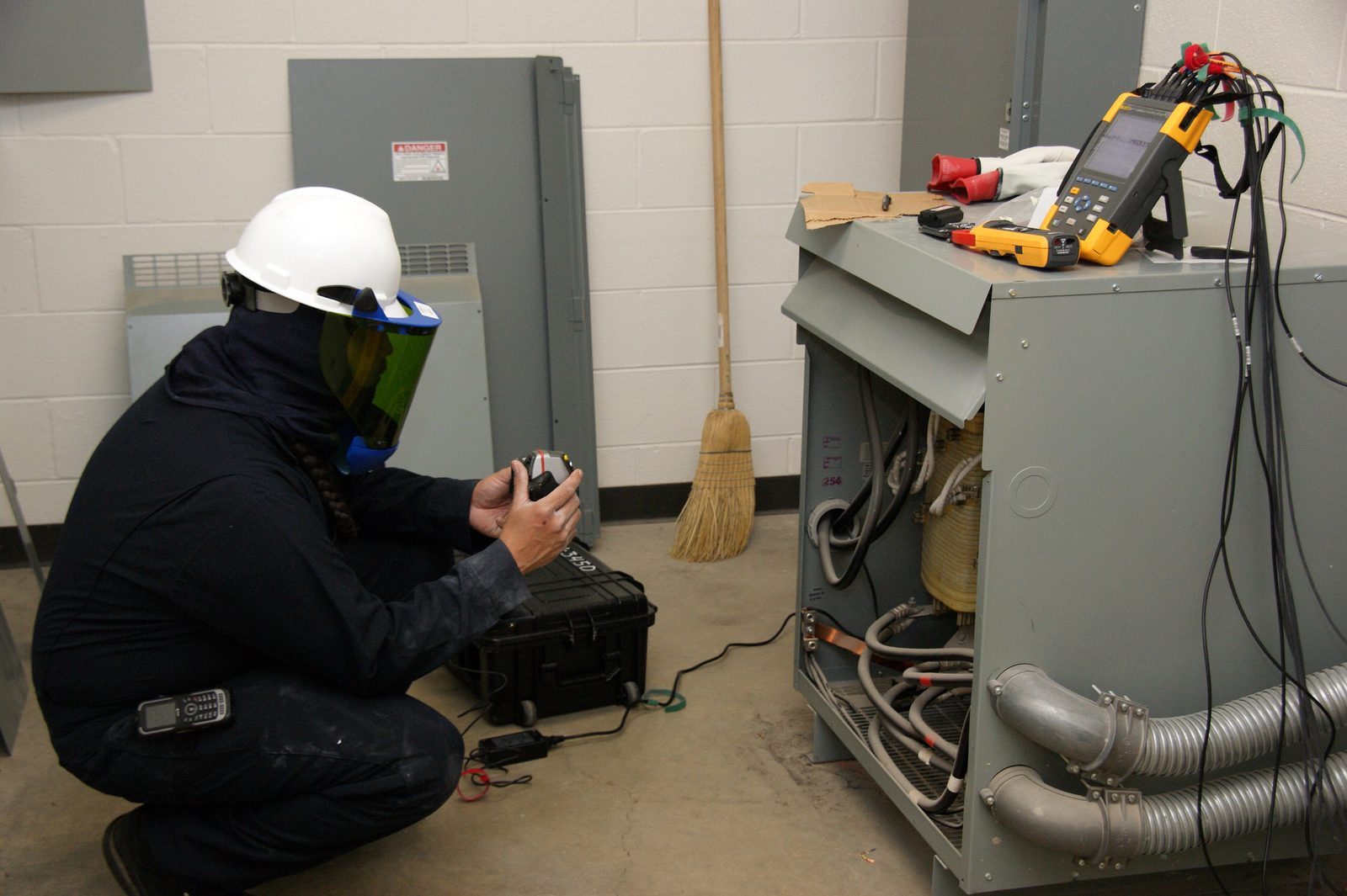 An electrician in a white hard hat and face shield crouches with a test instrument beside an open electrical cabinet with a Fluke meter on top.
