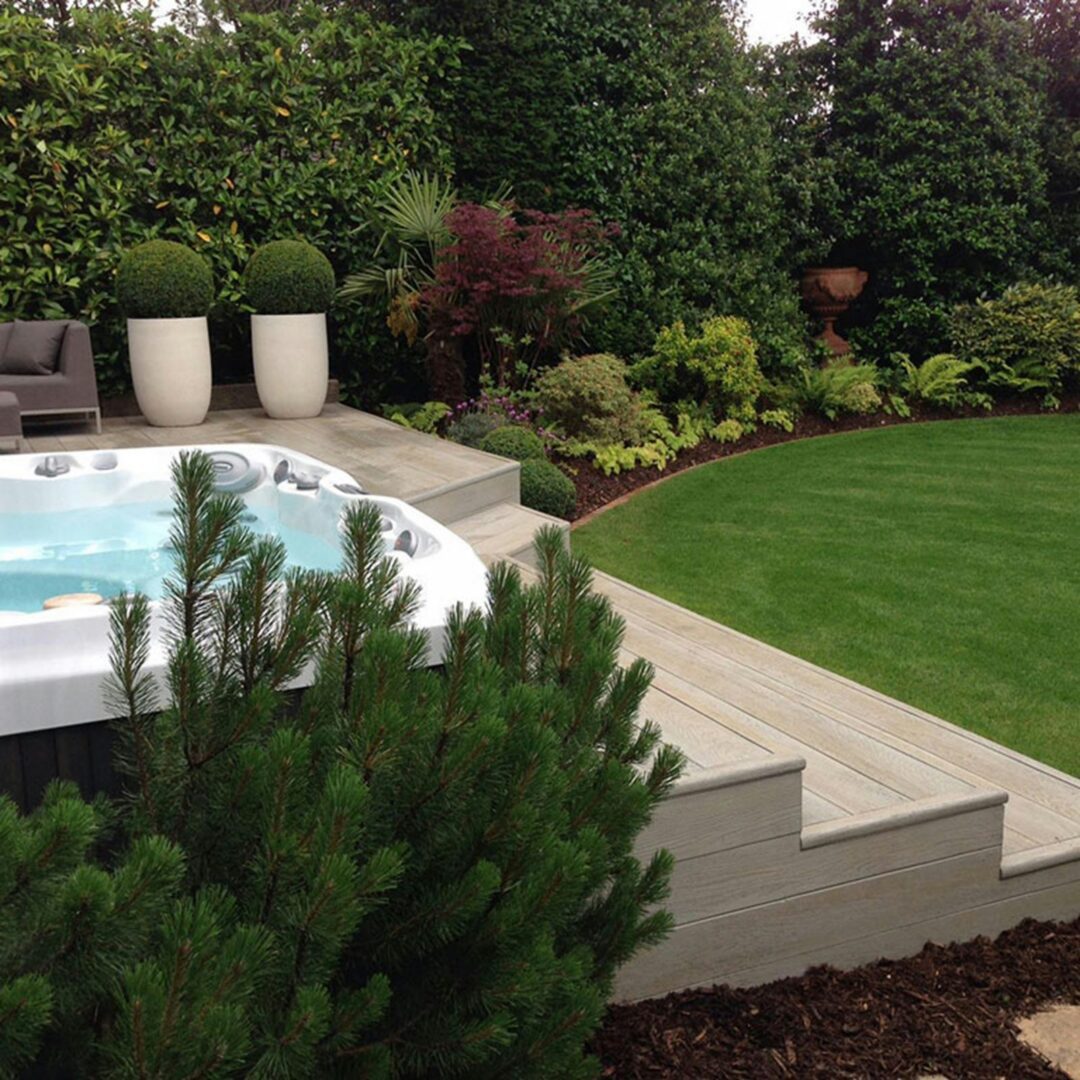 A white hot tub set into a wood deck beside a lawn and landscaped garden with potted topiaries.