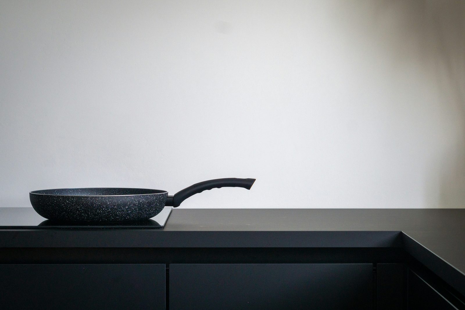 A black induction cooktop in a dark counter with a single frying pan on it against a white wall.
