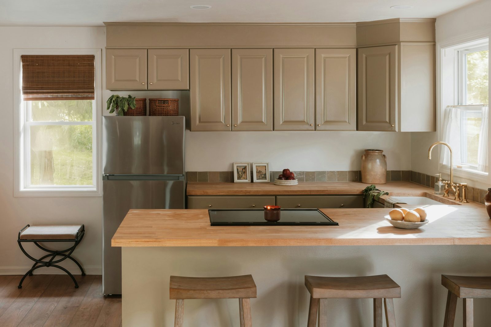 A warm kitchen with a butcher block island holding a black induction cooktop, stools and a stainless fridge.