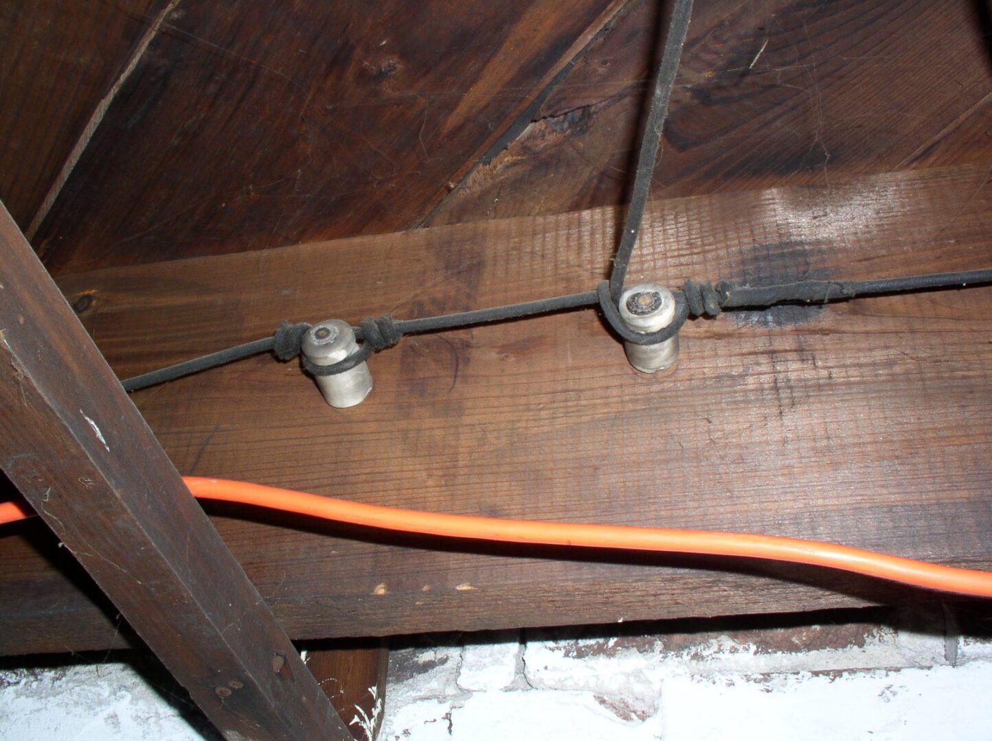 Two ceramic knobs supporting old wiring on a dark joist with an orange extension cord running beneath them.