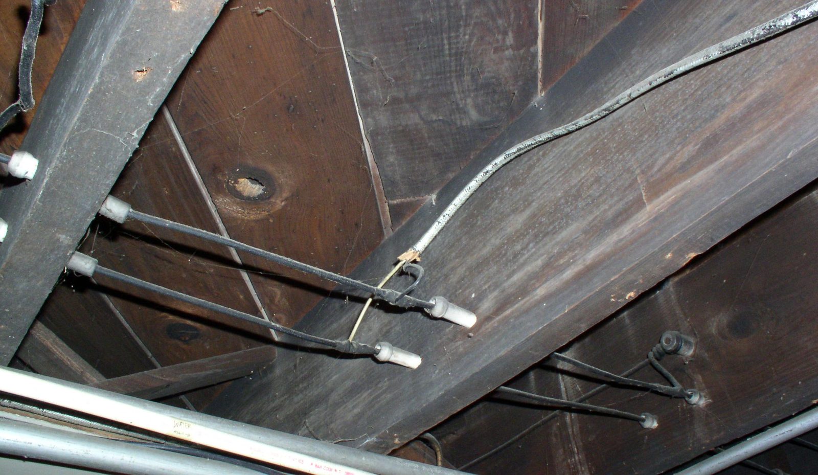Old knob and tube wiring running along dark wood floor joists with ceramic tubes passing through the framing.