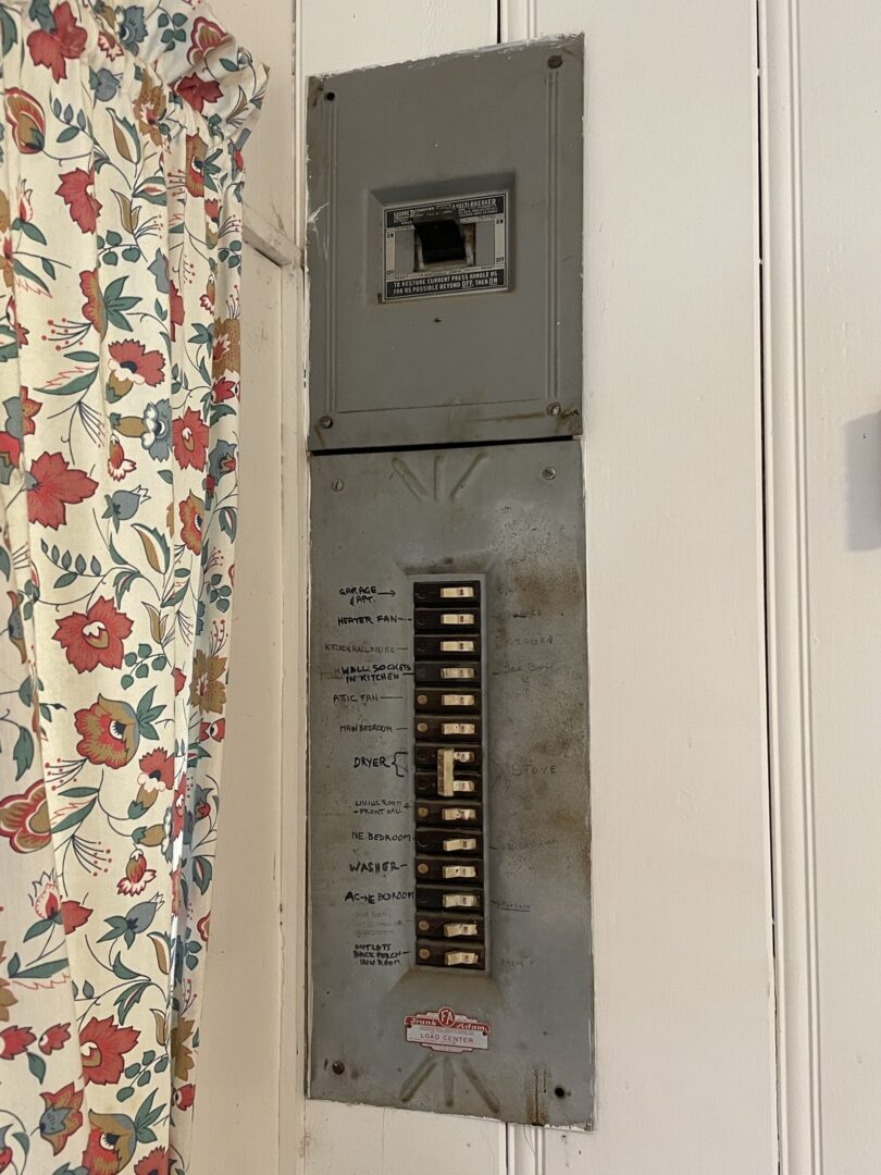 An old gray breaker panel set into a hallway wall with handwritten circuit labels next to each breaker, floral curtain alongside.