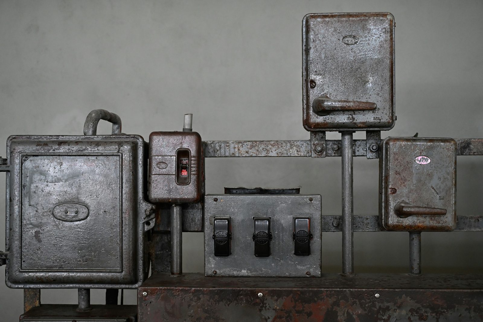 A row of old rusted gray metal electrical boxes and safety switches with lever handles mounted on a wall.