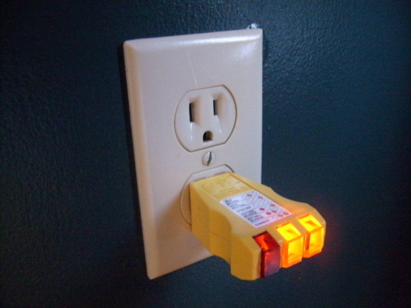 A yellow plug in outlet tester with lit indicator lights inserted in the lower outlet of a US duplex receptacle.