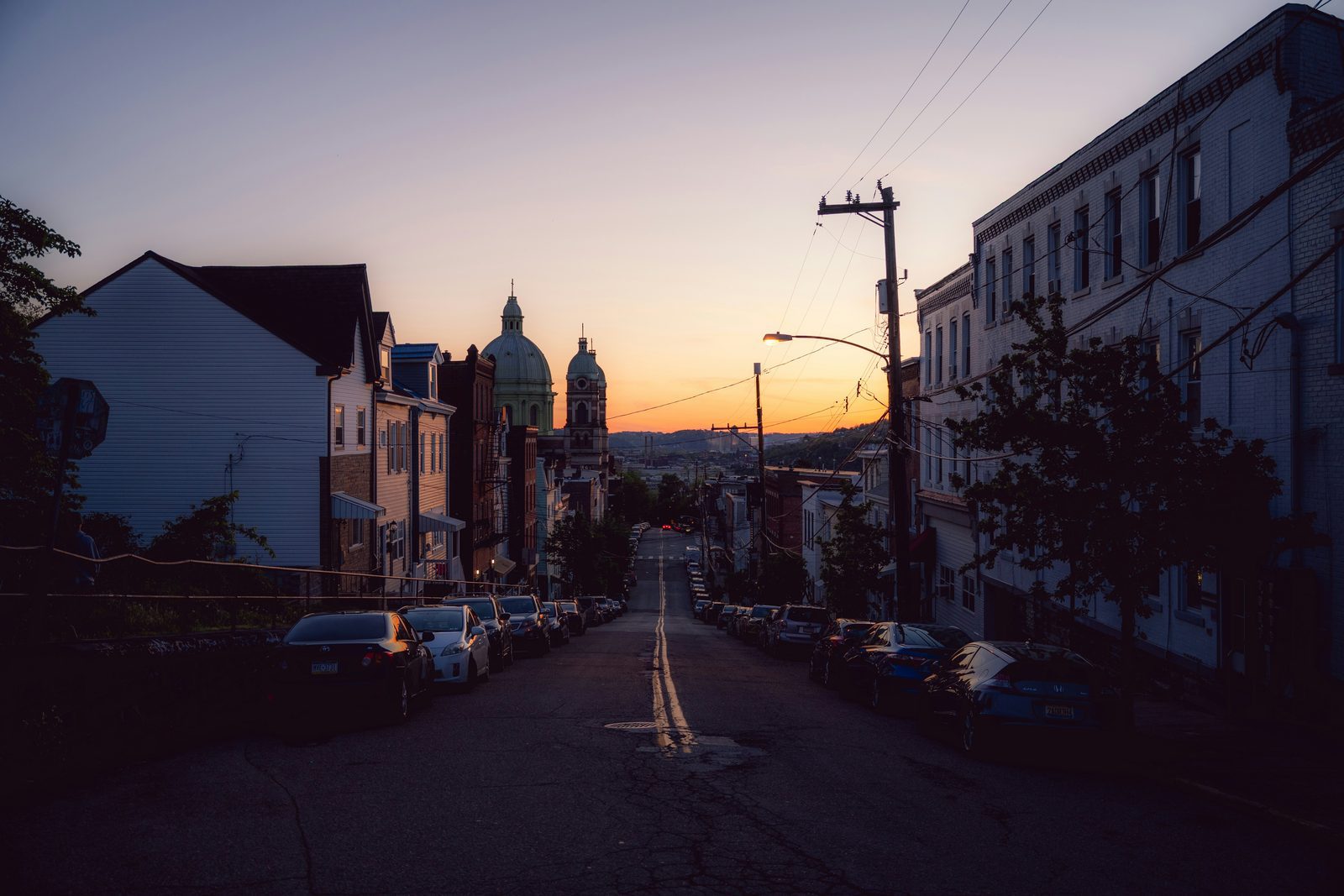 A Pittsburgh hillside street at sunset with parked cars, row houses and the domes of a church at the end of the street.