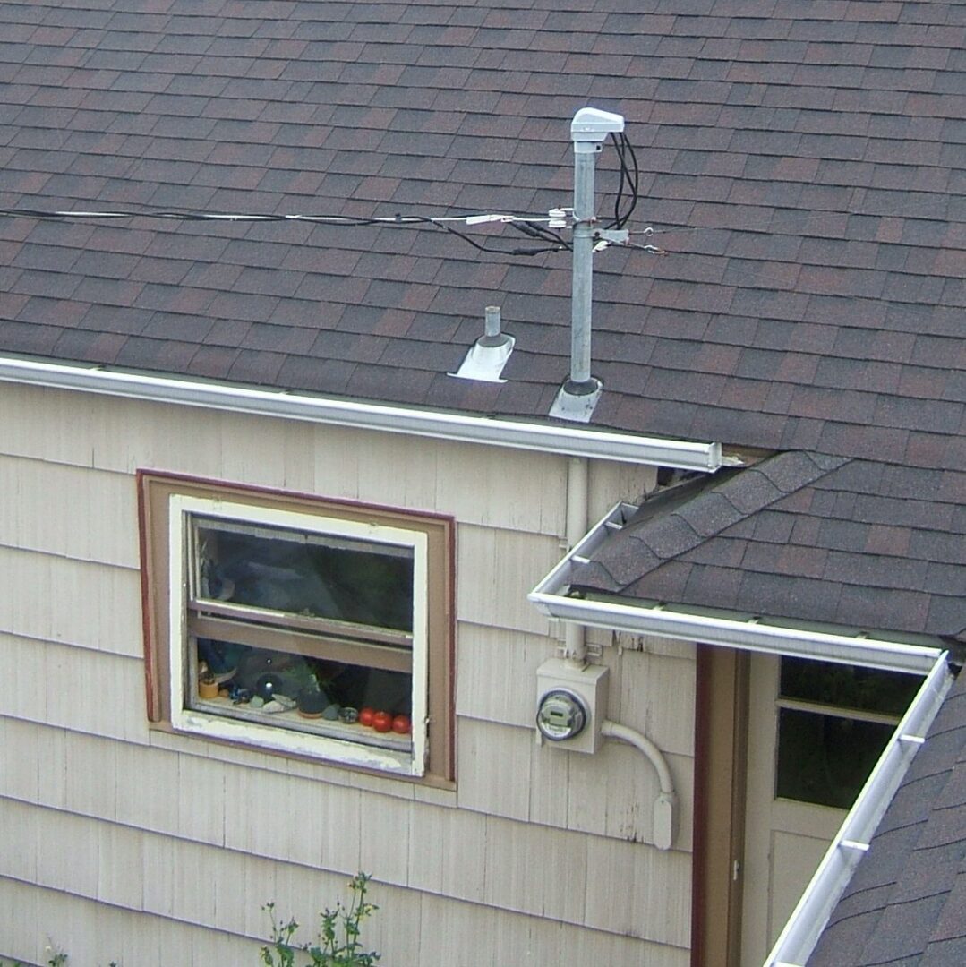 The roof corner of a shingle sided house with a steel service mast and weatherhead rising through the roof and the electric meter mounted below the window.