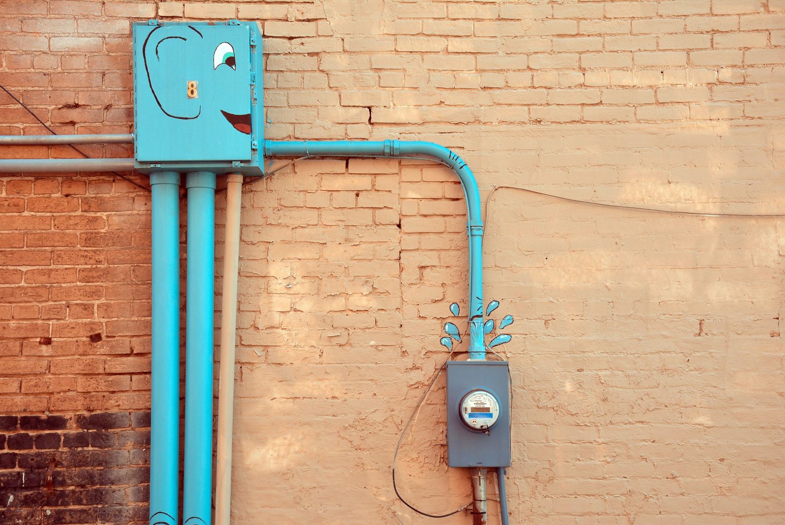 A single gray electric meter socket with round meter fed by teal painted conduit on a tan brick wall.