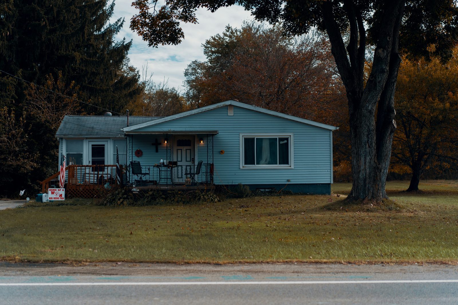 A small light blue single story house with a porch and an American flag beneath a large tree.