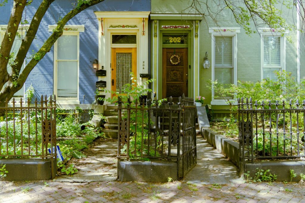 Two attached Victorian style row houses in blue and green behind an iron fence with overgrown front gardens.
