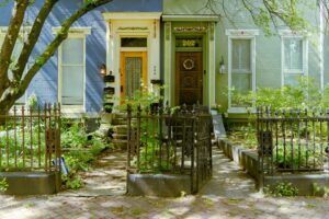 Two attached Victorian style row houses in blue and green behind an iron fence with overgrown front gardens.