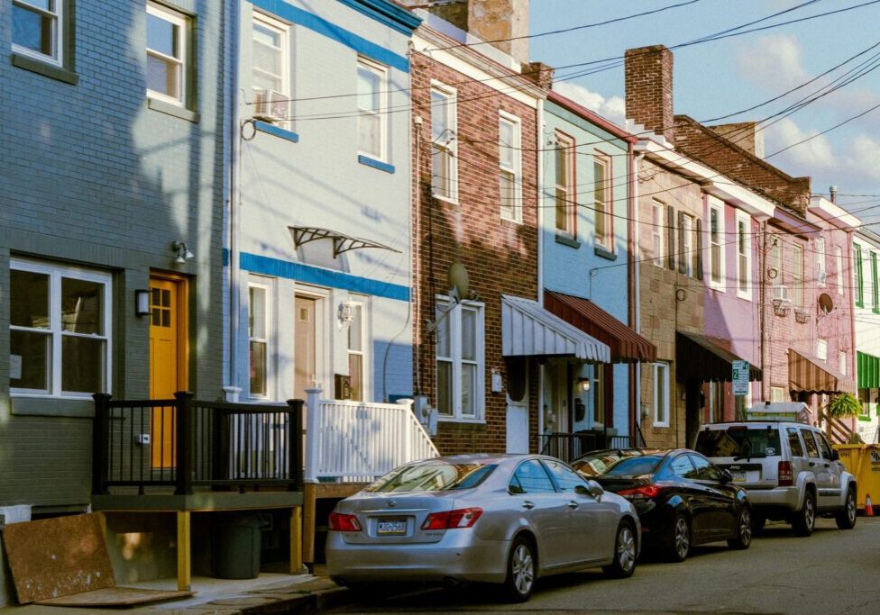 A row of attached two story brick row houses in blue, pink and tan with awnings and parked cars along the street.