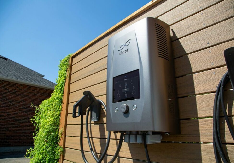 A silver dcbel home EV charger with a screen mounted on a wood fence outdoors, J1772 cable holstered beside it, brick houses in the background.