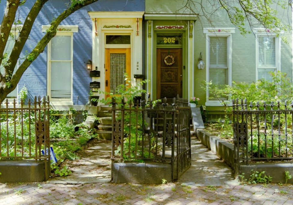 Two attached Victorian style row houses in blue and green behind an iron fence with overgrown front gardens.